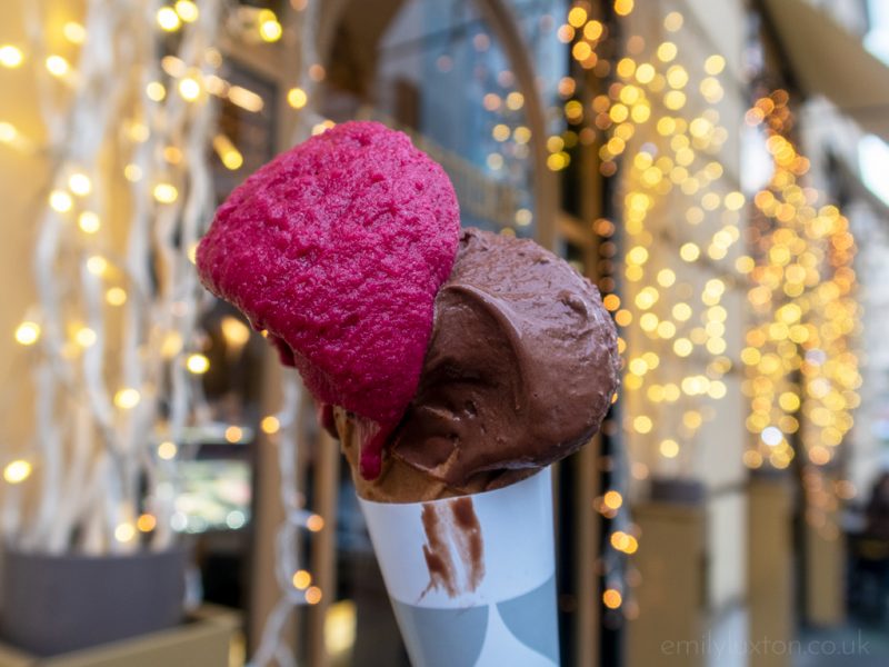 Close up of an ice cream cone with two scoops of gelate, one brown and one bright pink, in front of a shop covered with hundreds of fairy lights, at Neve di Latte Rome