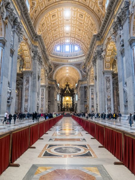 Interior of a St Peters Basilica in the Vatican with an arched cileing covered in golden tiles, the photo was taken on a quiet day in January and there is a small crowd of people walking through the cathedral