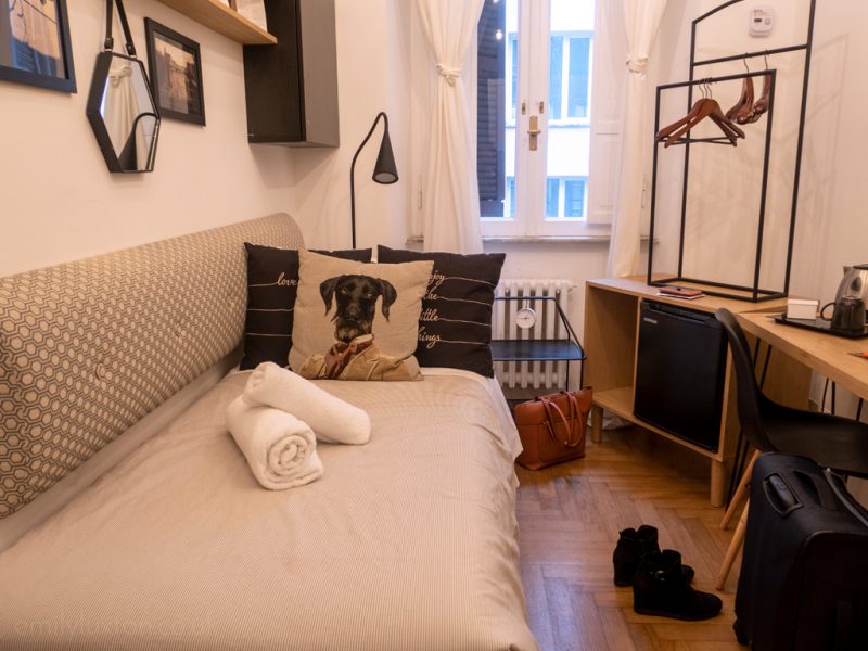 small hotel room with white walls and a single bed with white sheets and a cushion with a black dogs face on it