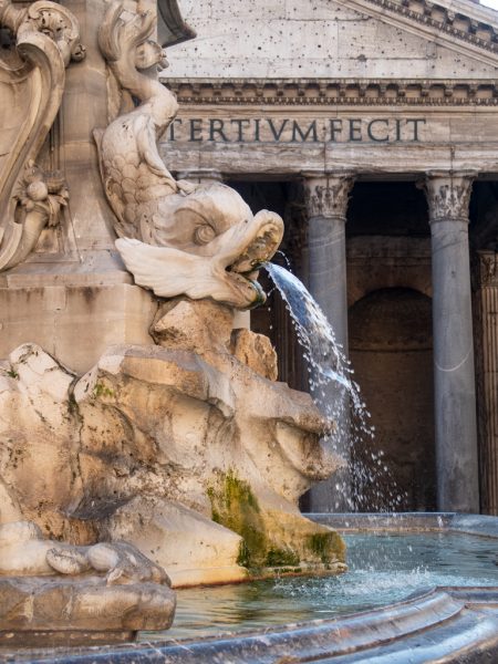 Close up of a fountain with a carved stone fish head spouting water in front of the stone pillars of the Pantheon in Rome 