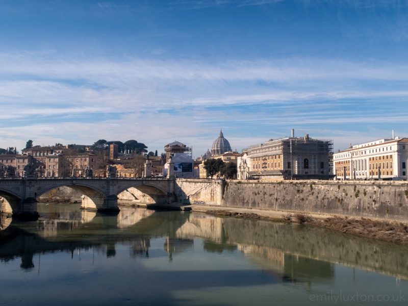 Wide, still river with a small three arched bridge across it and a city skyline on the other side. Rome in January