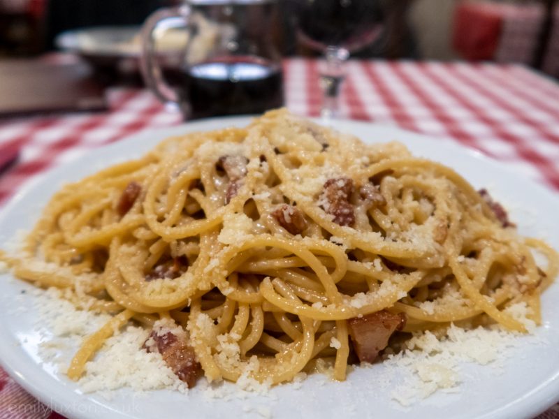 Close up of a plate of spaghetti with spall pieces of meal and lots of parmesan on a white and red checked tablecloth with a glass of wine behind