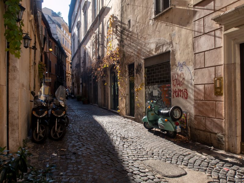 three mopeds in an alleyway with a cobbled street with late afternoon winter sun slanting between the buildings