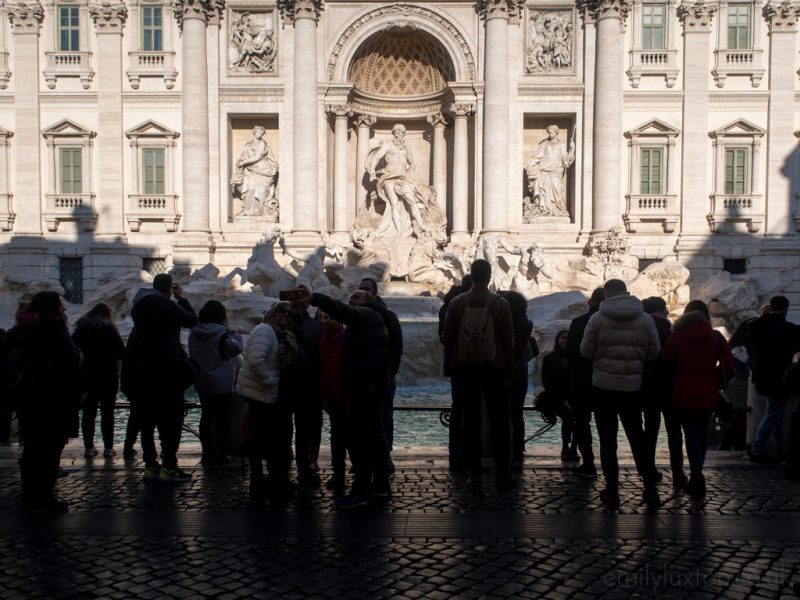 The Trevi fountain in Rome with a large white palace style builkding behind and a crowd of people silhouetted in front