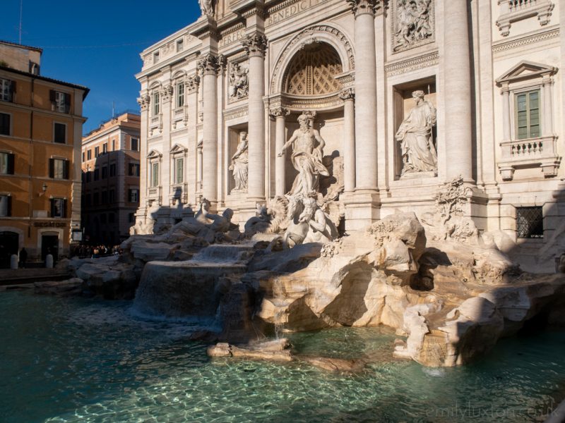 The Trevi Fountain with a large pool of cyan water overlooked by many white marble scultures of men and gods built in front of and into the facade of a white palace style building. Photographing Rome in January