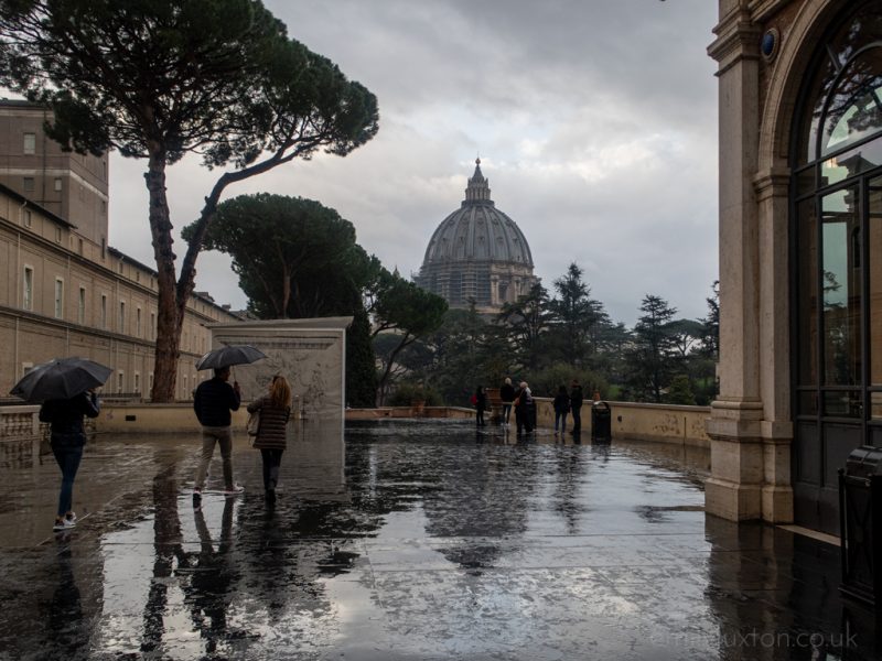 a cobbled square inside the Vatican on a rainy day with wet floor and the dome of St. Peter's Basilica above some trees against a grey sky