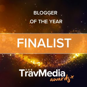 square graphic with a dark brown background with yellow and gold sparkles, the text in white reads: blogger of the year finalist The TravMedia awards"