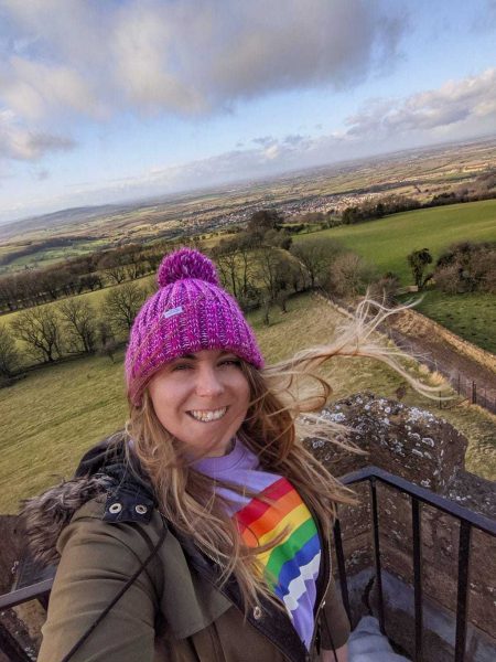 Selfie of Emily wearing a purple jumper wiht a rainbow on it, a khaki parka coat with a fur lined hood, and a pink wooly hat over her long blonde hair which is blowing in the wind. she is standing by a stone wall on top of a tower with green fields and trees below in the Cotswolds countryside.