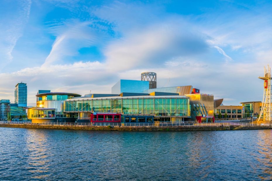 River with a city skyline on the far side with several waterfront buildings mostly made of glass. It is a very sunny day with blue sky above. places to visit in the north of england - manchester
