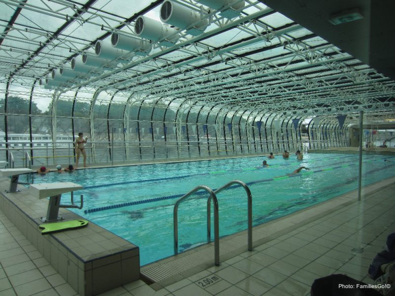 Indoor simming pool with a glass rood and wall. the pool is set up for lane swimming and there are a few people swimming in the far end. a woman in a red bikini is standing by the glass wall on the far side of the pool. unusual things to do in Paris