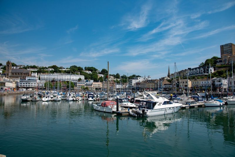 Marina with numerous boats docked in calm water, surrounded by residential and commercial buildings. Clear sky with a few wispy clouds, creating a picturesque coastal scene on a sunny day.