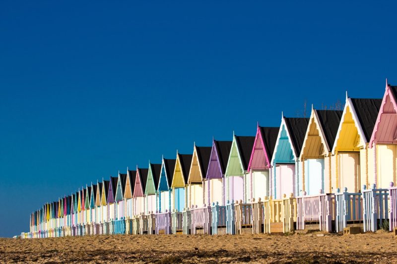 Row of wooden beaches with white fronts and brightly painted roofs, each one painted a different colour, on a shingle beach in Mersea Island Essex on a very sunny day with clear blue sky above