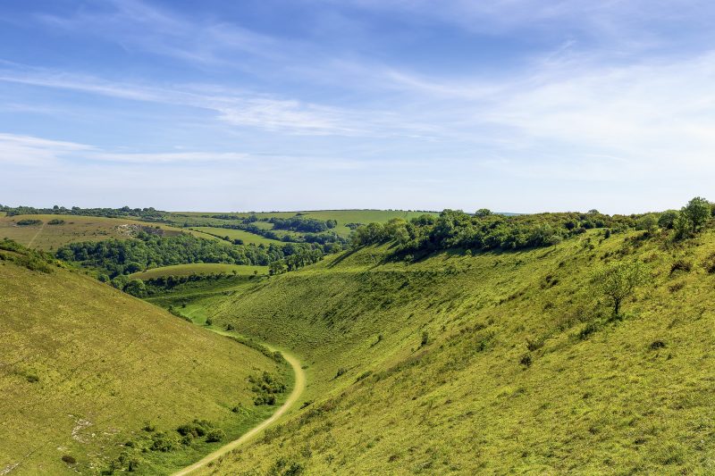 Footpath in a grassy valley in the South Downs in Hampshire England, with grassy slopes on either side and green countryside in the distance, on a sunny summer day with blue sky above.