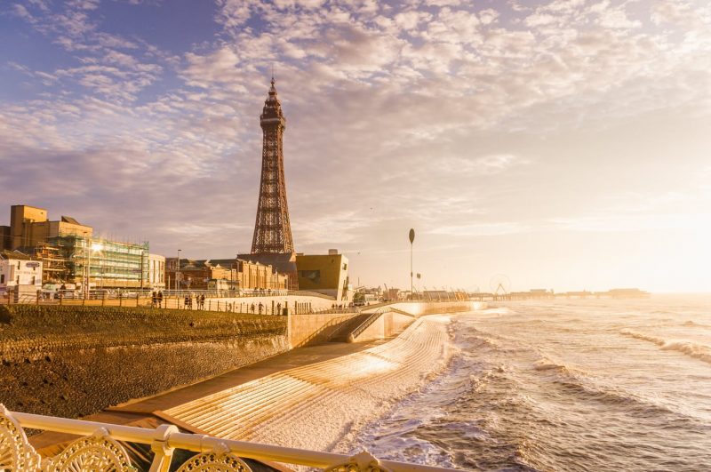 Blackpool seaside in the North of England, with a metal tower on the esplanade to the left overlooking the sea on the right, taken near sunset with the sun going down over the sea in the far right of the picture.