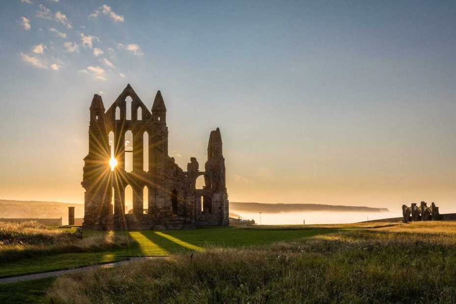 Ruins of Whitby Abbbey at sunset, a stone wall with tall arched windows in it with the setting sun flaring through one of the windows. The abbey is on a grassy clifftop with a misty bay beyond and a low headland beyond that with clear blue and yellow sky overhead. Best Places to Visit in the North of England