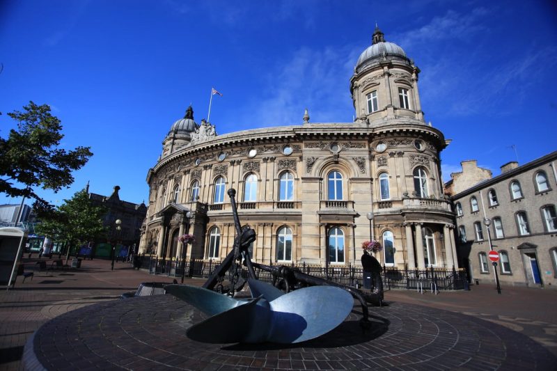 Exterior of a semi circular shaped two-storey building of light grey stone with arched windows and a domed turret on the left hand side. It is a very sunny day with clear blue sky behind. Kingston Upon Hull, Northern England
