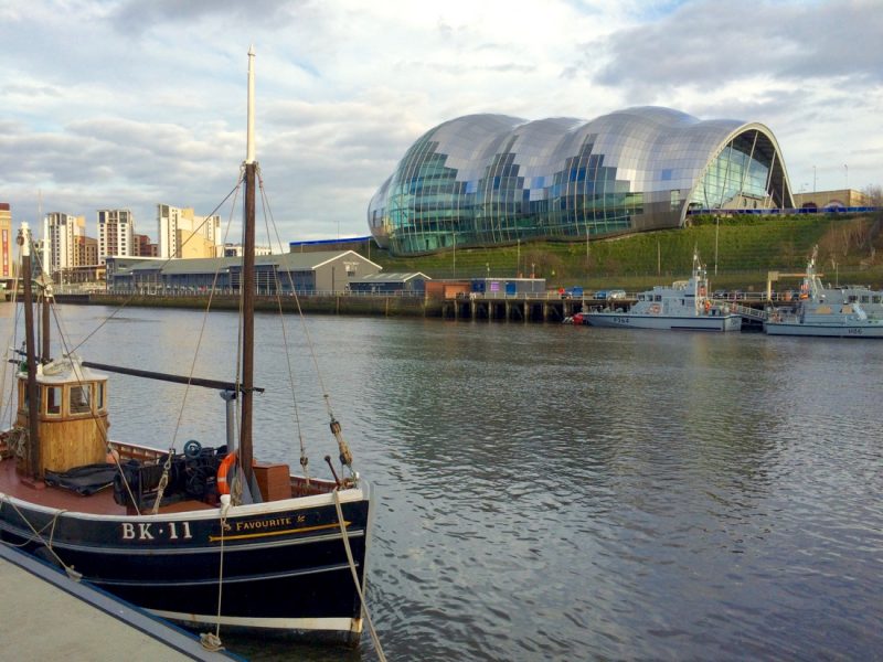 Wide grey river with a blue wooden fishing boat in the foreground and a large bubble shapred glass building on the far side of the river in Newcastle. Best cities to visit in northern england