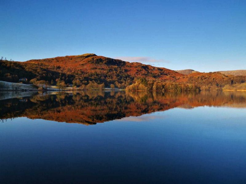 Looking across a large flat lake with very blue water towards a low hill covered with red and yellow trees on a sunny autumn day with clear blue sky and the hill perfectly reflected in the lake. Lake District England in Autumn