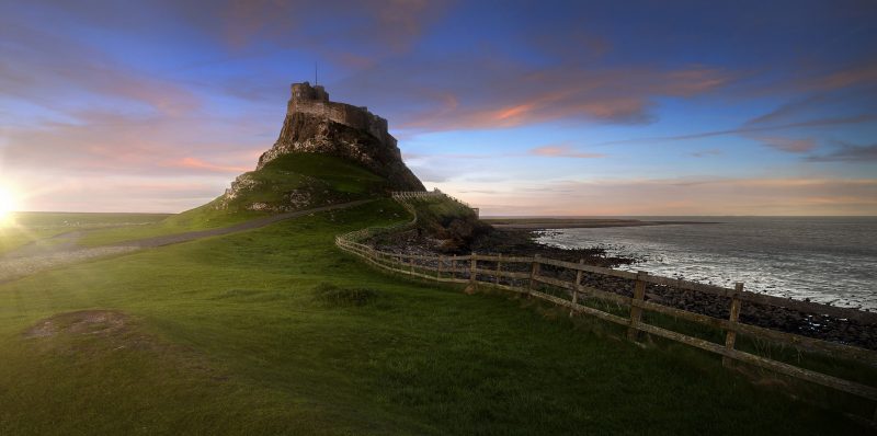 Castle with a circular stone wall around it on the top of a small rovky hill on Lindisfarne Island with the sea on the right and a large empty area of grass in front and to the left. Best Places to Visit in the North of England