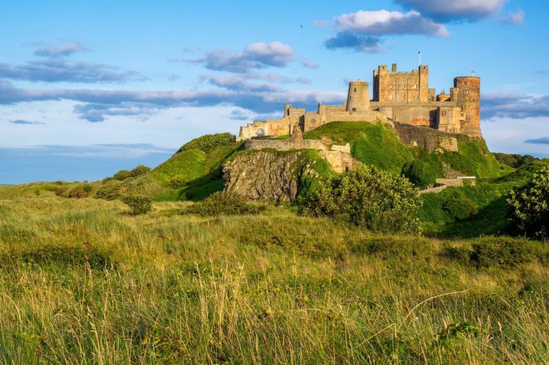 Bamburgh Castle in North England, a light grey stone castle at the top of a smlal grassy hill with long grass in the foreground, taken on a sunny day with bright blue sky and small clouds above.