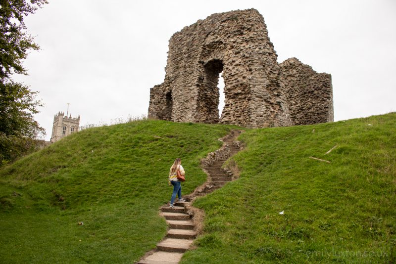 Emily wearing blue jeans and a white and yellow striped wooly jumper with her blonde hair down and carrying an orange handbag. she is walking up a stone staircase on the side of a grassy hill towards a ruined stone castle with overcast sky overhead. Autumn Packing List for England - City break