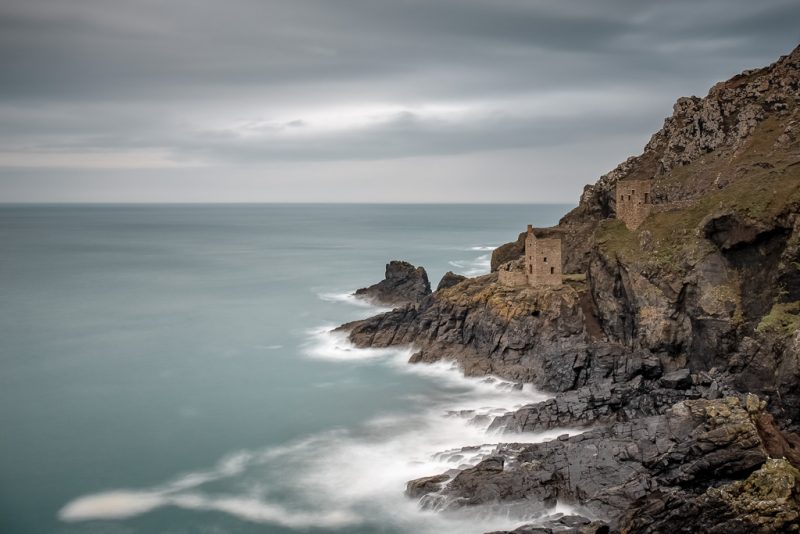 Looking across a section of the North Cornwall Coast in England in Autumn with the sea a very dark greyish blue and the sky overcast. There is a small, rocky cliff with jagged grey rocks at the bottom. there are two small stone buildings, partly ruined and without roofs, on the sloping edge of the cliff over the sea. 