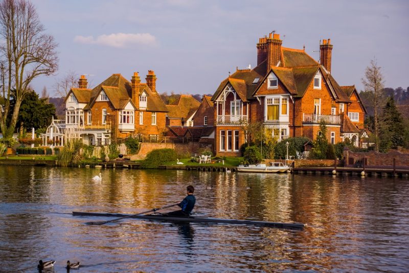 Man rowing a thin grey rowboat along the river in Marlow buckinghamshire near some swans. there are two large red brick houses on the far bank and a small white boat parked at the edge of the river. 