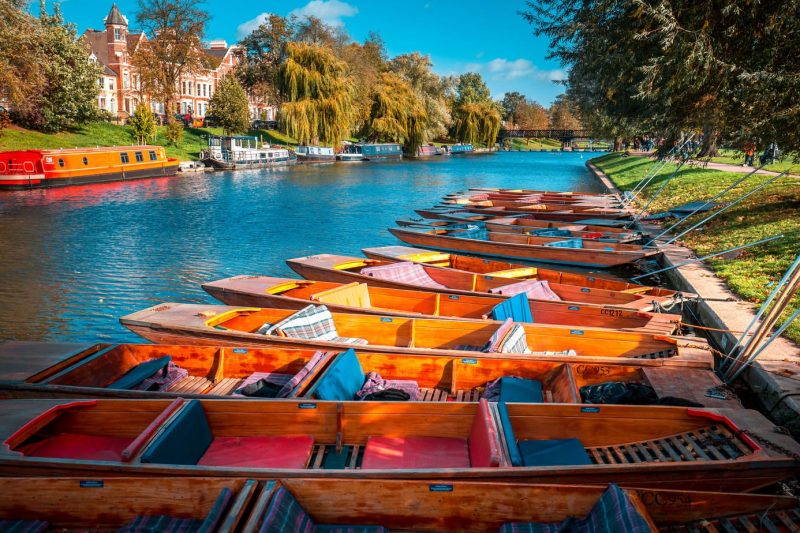 A row of wooden rowing boats in the river in Cambridge in Autumn with trees on either side of the river and an orange canalboat on the far side with a row of red brich terraced houses visible through the trees behind that. 