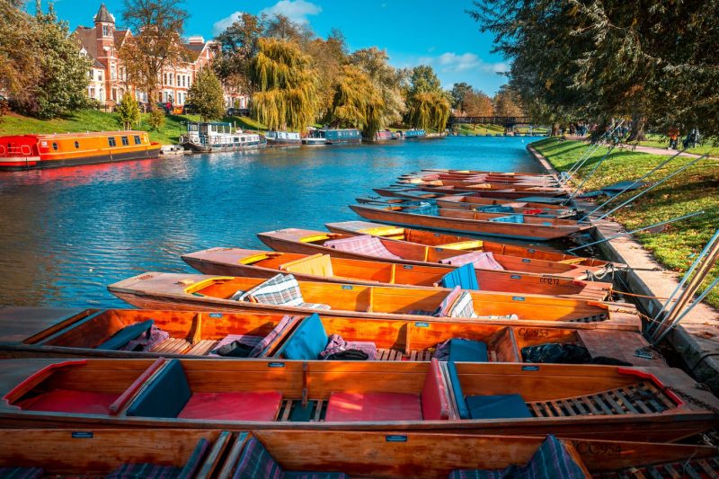 A row of wooden rowing boats in the river in Cambridge in Autumn with trees on either side of the river and an orange canalboat on the far side with a row of red brich terraced houses visible through the trees behind that.