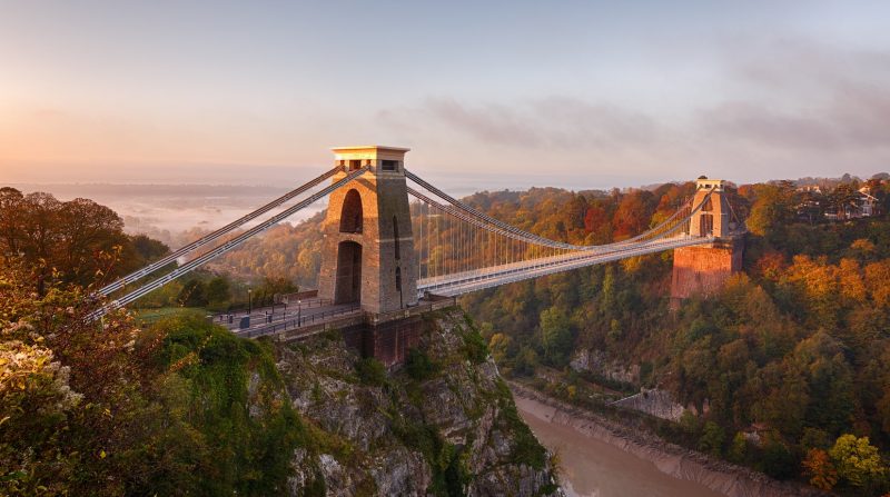 Looking down at Clifton Suspension Bridge above the River Avon in Bristol, the bridge has a stone tower on either side with long grey metal cables between each one holding up the bridge. the banks fo the river are covered with a dense forest with green and golden leaves. 