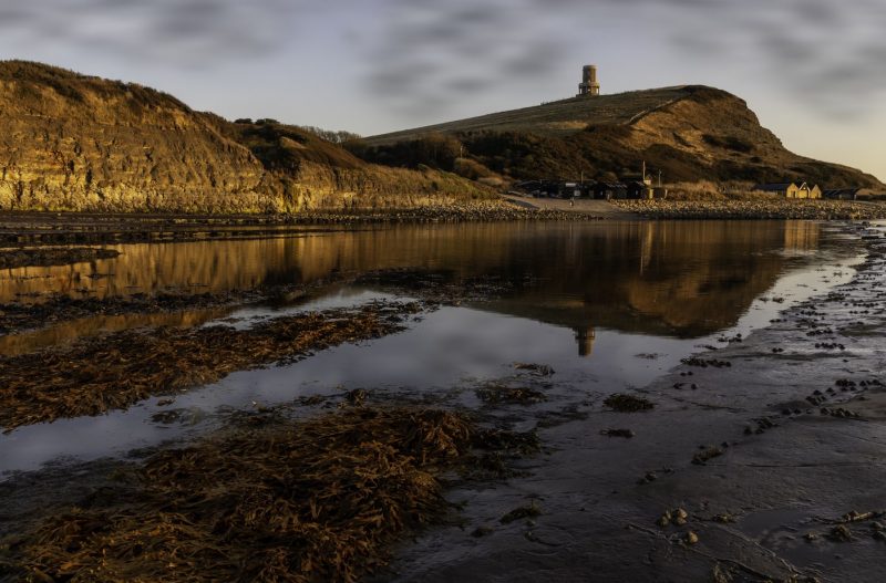Kimmeridge Bay in Dorset with a small stone circular tower on top of a low grassy headland above a small shingle and rocky beach. The tower and headland are perfectly reflected in the water in the way. 