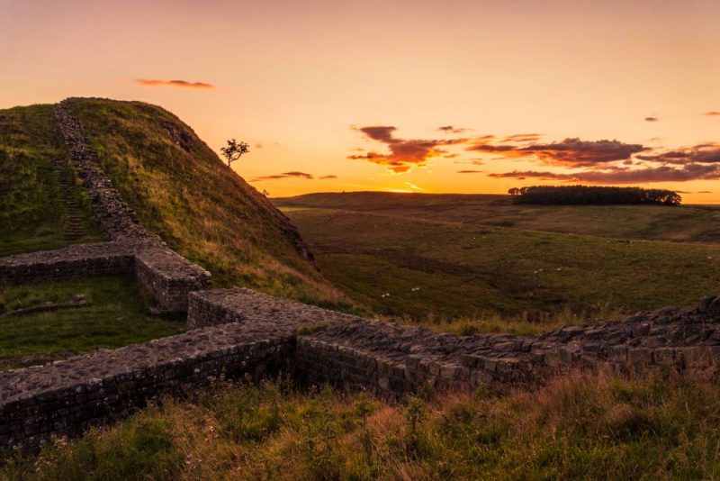 Section of Hadrians Wall in England taken at sunset in autumn. there are parts of an old ruined wall visible running uphill with a wide grassy valley beyond and the sun setting over the hill on the far side with a small woodland silhouetted against the orange sky. 