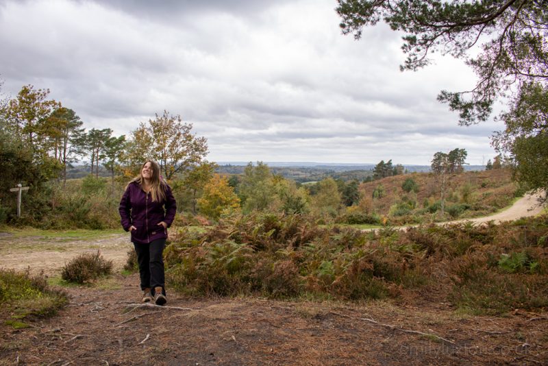 Emily wearing a purple down raincoat, black walking trousers and brown hiking boots with her long blonde hair down and her hands in her pockets walking towards the camera and smiling on a dirt path. there is a gravel path behind her and a heath behind that on an overcast day with grey sky above and reddish brown ferns all around the path emily is walking on. Autumn Packing List for England.