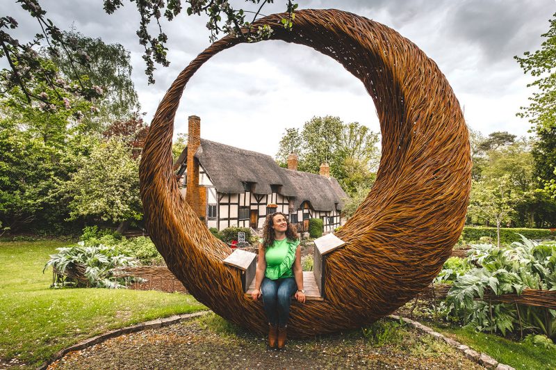 A lady wieht very curly brown hair wearing blue jeans and a bright green vest top with a ruched detail across the front, sitting in a wooden seat inside a circular wicker structure, with a thatched roof cottage with cream half timbered walls behind in Stratford-upon-Avon