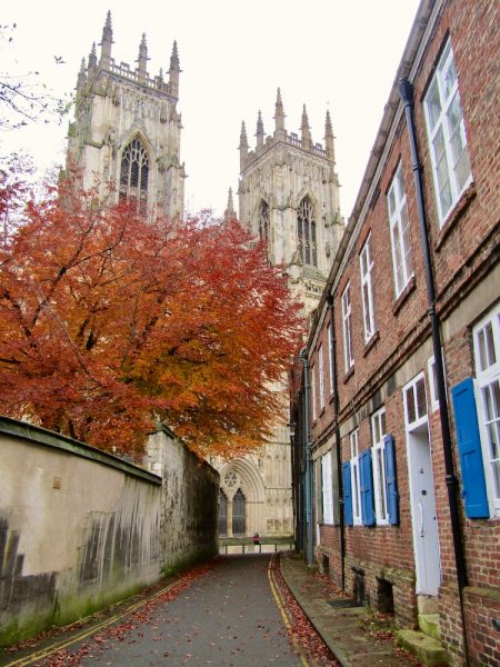 The towers of York Minster appearing above red Autumn Leaves at the end of a narrow alleyway