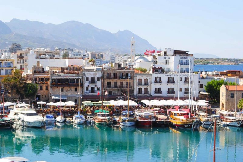 A picturesque harbor scene with boats, a mix of rustic buildings, hotel signs, and mountains under a clear sky.