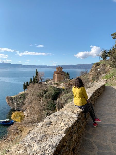 A person with bob-length black hair in a yellow jacket and pink running shoes sits on a stone wall, overlooking a calm lake with a historic building on a cliff.