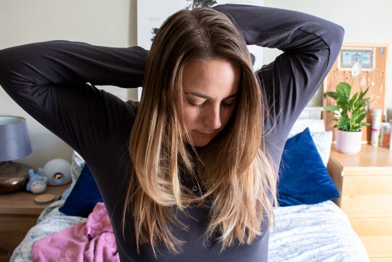 emily wearing a grey long sleeved top with her arms behind her head stretching while she looks down with a white double bed behind her