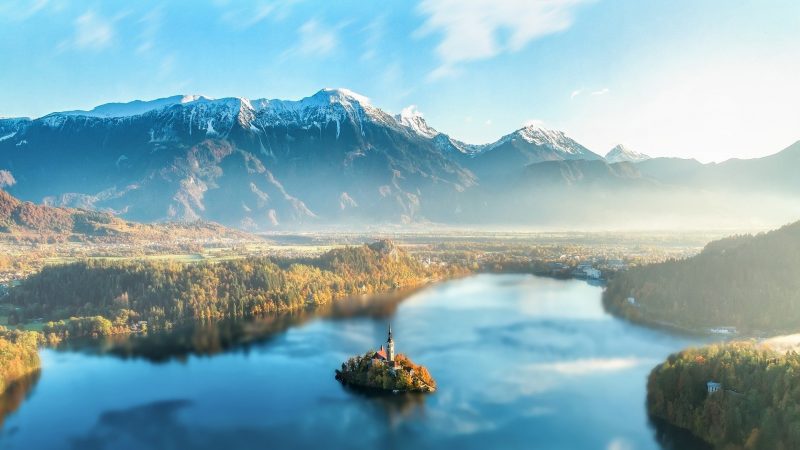 A picturesque landscape featuring a serene lake with an island in the center, autumn trees, and a church steeple surrounded by lush greenery, with snow-capped mountains in the background under a clear blue sky.