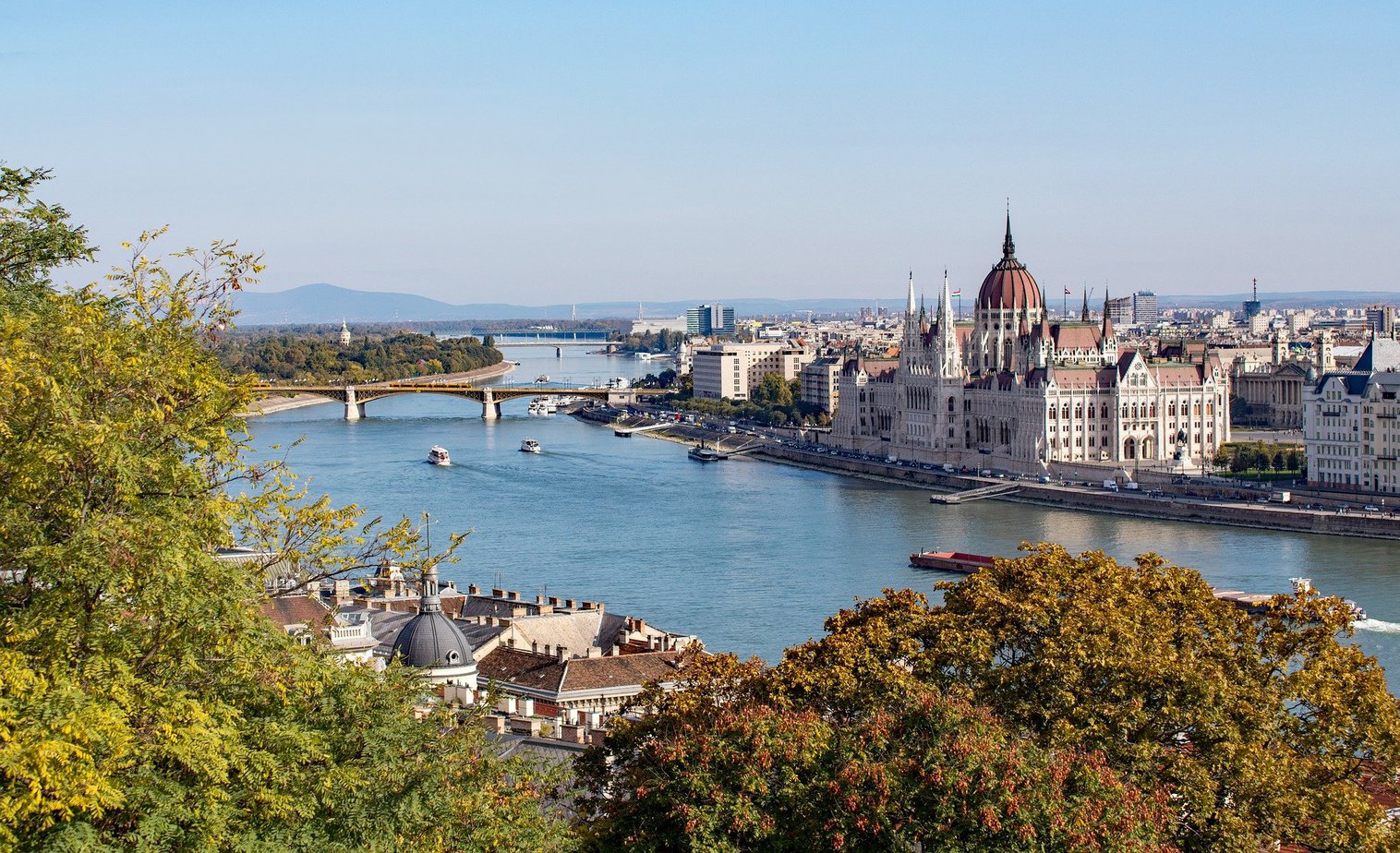 View from a hillside of the Danube River in Budapest with a bridge across the river in the distance and a large palace style building on the far bank with white stone facade and a red domed roof. Best river cruises in europe