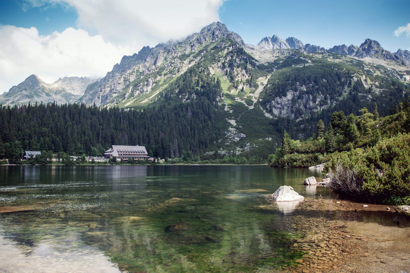 A serene lake surrounded by lush greenery and towering mountains, with a gabled-roof building near the lake's edge.