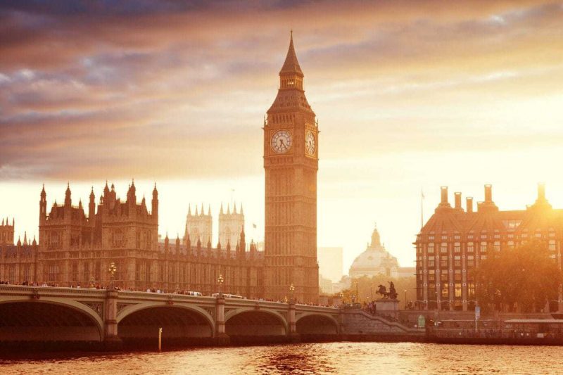 Big Ben and the houses of Parliament in London with the River Thames in front at sunset, the sky behind is yellowy-gold and the flat river is reflecting the sunset light. most beautiful cities in england. 