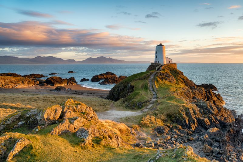 rocky coastal landscape with a small white lighthouse standing on a rocky hill near the sea and a rock path leading down from the grassy mainland on a very sunny day in Anglesey - best road trips in Wales