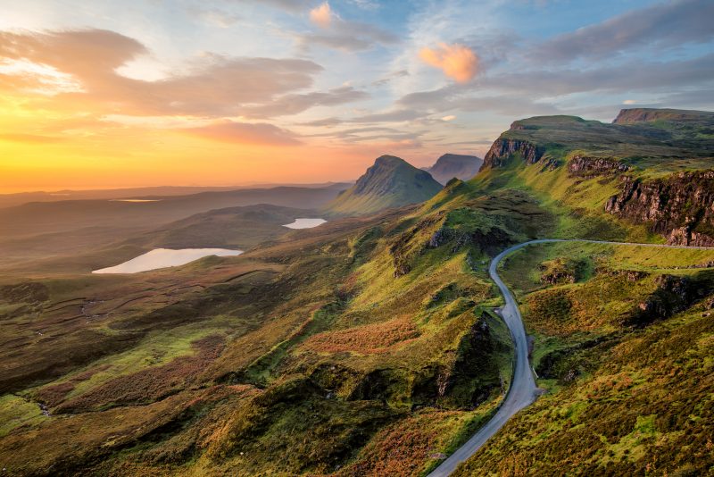 Winding road on the Isle of Skye on a graassy hillside with a dramatic landscape of peaks and lakes visible behind at sunset with an orange sky. UK Staycation Ideas - Scottish Islands