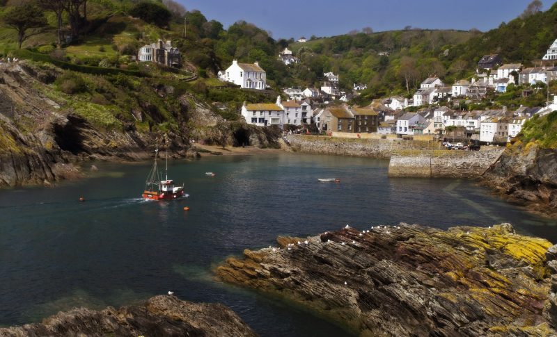 Looking acoss Polperro Harbour in Cornwall with a red and white fishing boat approaching the harbour and the small village of white houses clustered along the shore