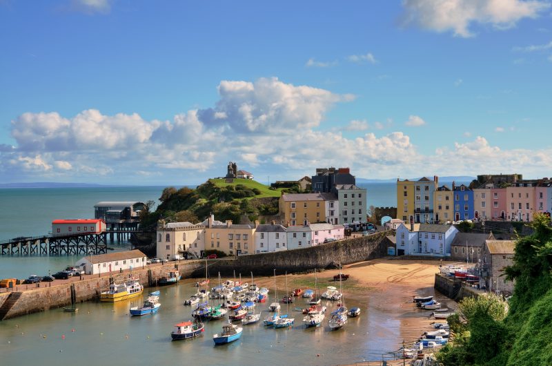 small harbour filled with colourful fishing boats with a small sandy beach behind and a town full of colourful houses in different pastel shades behind that. there is a small green grassy hill in the background and the blue sea visible behind it all on a vwery sunny day with blue sky overhead. 