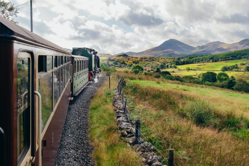 steam train with a dark green engine and dark crimson carriages running through snowdonia with mountains in the distance - the photo is taken out the window of the train looking along the carriages towards the mountains 