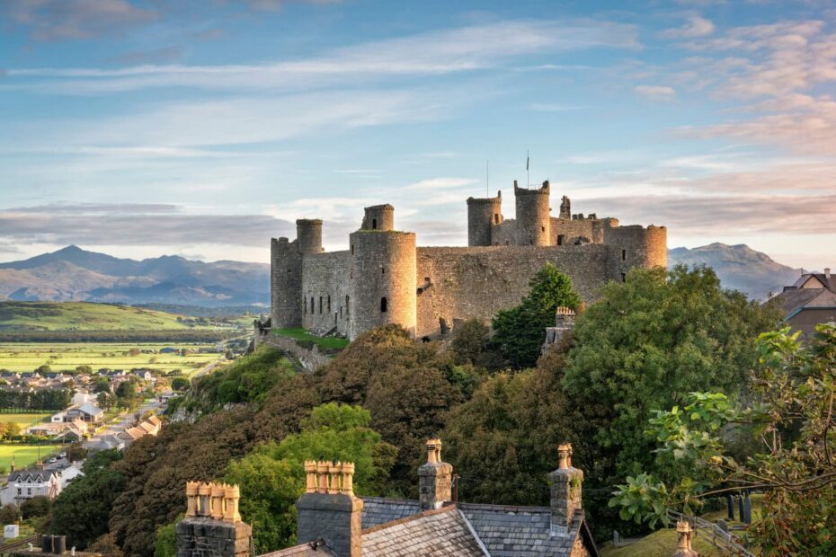 The historic Harlech castle on a hill with multiple towers and battlements overlooks a small town. The scene is lush with green trees and vegetation, fields, and distant mountains under a partly cloudy sky. Houses with chimneys are visible in the foreground, and it is just after sunrise with the warm lighting.