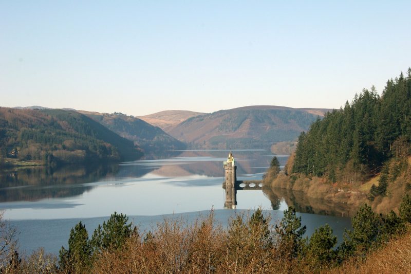 Lake Vyrnwy in North Wales with a large blue flat lake surrounded by low mountains and pine trees. there is a small stone tower on a short bridge jutting out from the land to the right.