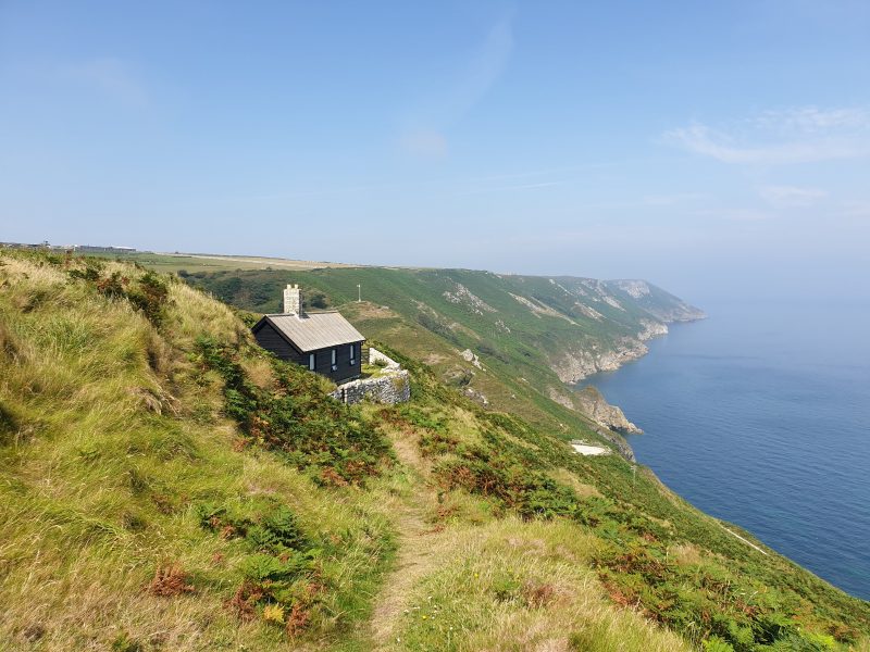 Looking along the grassy cliffs of the Lundy Island coastline with the sea on the right and a small stone cottage tucked against a grassy bank near the cliff edge